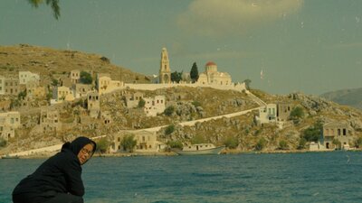 Woman sits on edge of water bank wearing black garment, mountains in background