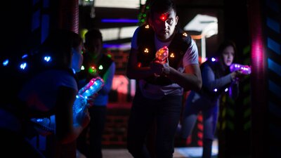 Players in neon-lit laser tag arena wearing glowing vests and aiming light-up laser guns
