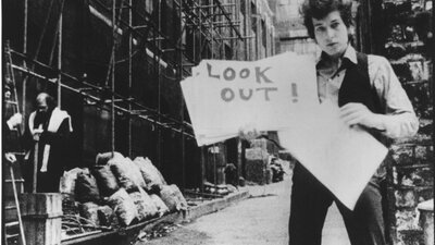 Black and white photo of Bob Dylan holding a poster that says look out!