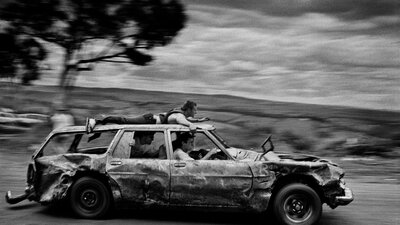 black & white image of a banged-up car speeding past with passengers and one man on the car roof