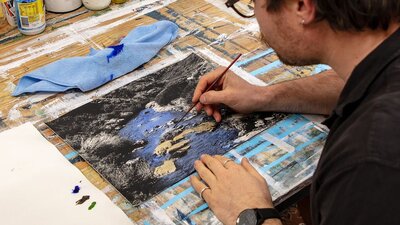 A photo of a man painting a black and white darkroom image of a river