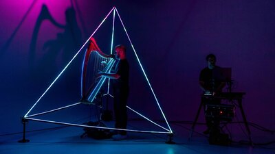 A harpist performs inside a glowing triangular light structure, operated by a man in the shadows