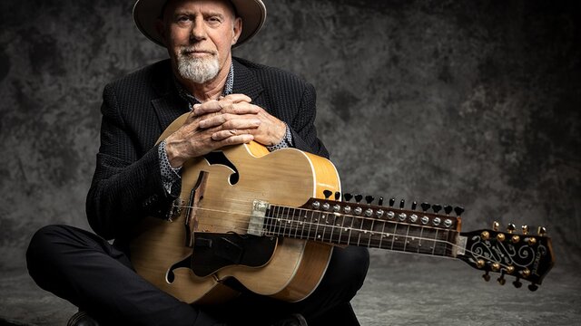 Harry Manx sitting down, holding stringed instrument