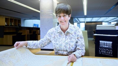 A smiling woman in a floral shirt sits at a desk, holding open a large historical map.