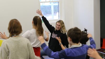 A young woman kneels and extends an arm in a dramatic gesture. A group of children imitate her.