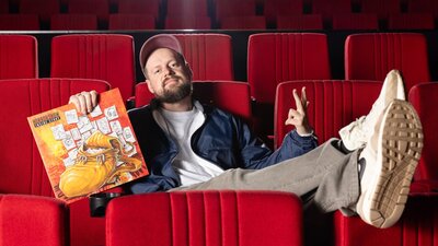 Horrorshow x CLIPPED: Inside Story Uncut (2025) Man sitting in an empty cinema, feet up on seats with a cap, holding a vinyl cover