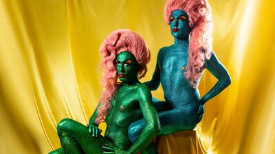 Two figures with flowing hair and sparkly skin sit together elegantly, a gold sheet draped behind.