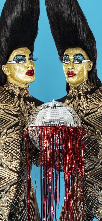 Two figures with tall hair and extravagant, pointed outfits hold a disco ball with red tinsel.
