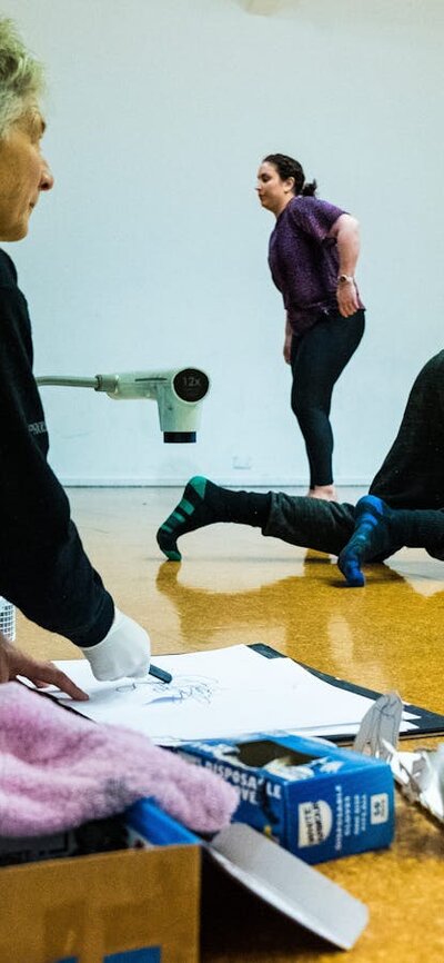 2 figures in the background are dancing. The woman in the front kneels next to various art supplies.