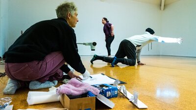2 figures in the background are dancing. The woman in the front kneels next to various art supplies.