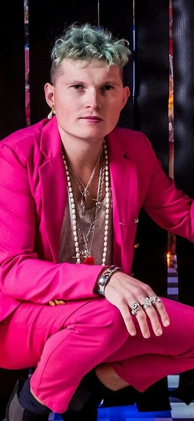 An adult wearing a pink suit poses on the ground in a colourful, decorated room.