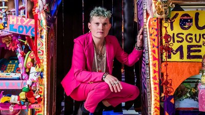 An adult wearing a pink suit poses on the ground in a colourful, decorated room.