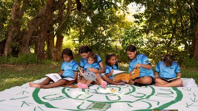 Image courtesy of the Indigenous Literacy Foundation. A group of diverse young children are sitting outdoors under lush green trees, engaged in reading.