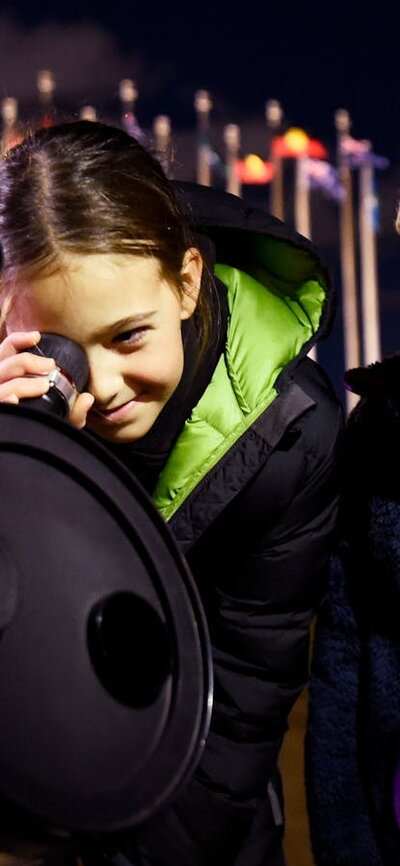 Two children stand outdoors at night using a telescope.