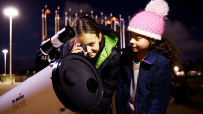 Two children stand outdoors at night using a telescope.