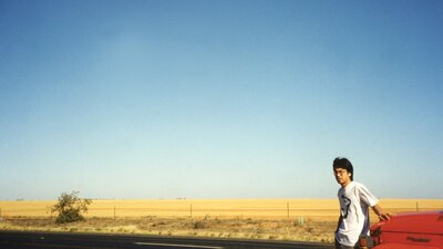 Photo of man standing on the right leaning on a car with a desert in the background and blue skies