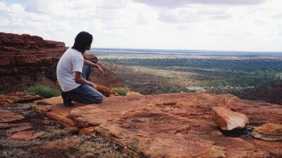 Man kneeling on red dirt on a cliff, looking into the distance