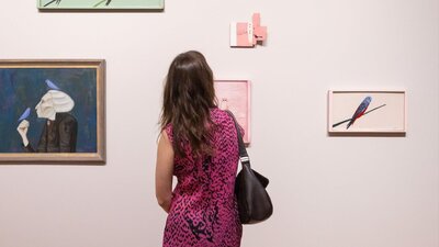 A person wearing a patterned dress views several painted portraits on a light-coloured wall.