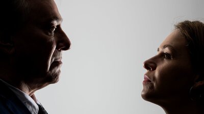 A man in a suit and a woman in a blouse look intensely at one another.