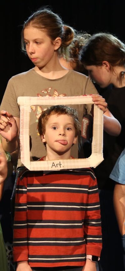 A group of primary school-aged children stand together on a dimly lit stage, gathered closely as if Children on stage create a playful tableau around a child framed by a white picture frame labelled “