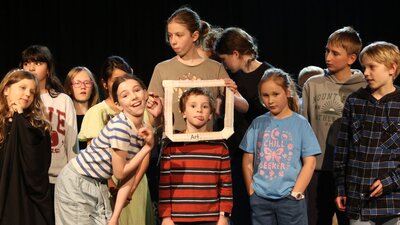 A group of primary school-aged children stand together on a dimly lit stage, gathered closely as if Children on stage create a playful tableau around a child framed by a white picture frame labelled “