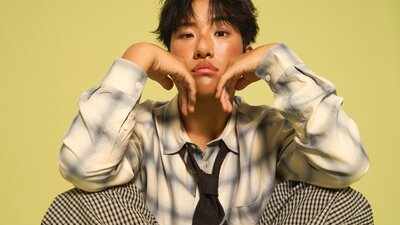 KINKY sitting down with his hands placed under his chin, with a yellow background