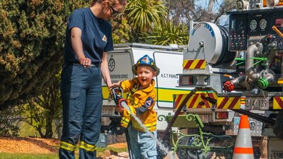 A child using a firehose while supervised by a volunteer firefighter