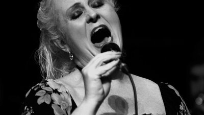 Black and white photo of a woman singing into a microphone wearing a floral dress