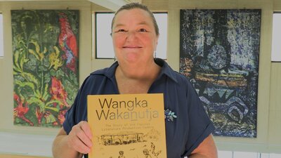 A woman holding a book with a yellow cover, standing in front of some wall tapestries