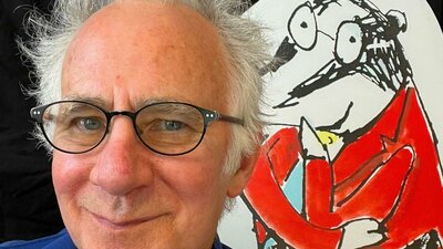 Man with glasses smiles beside a cartoon bear in a red jacket.
