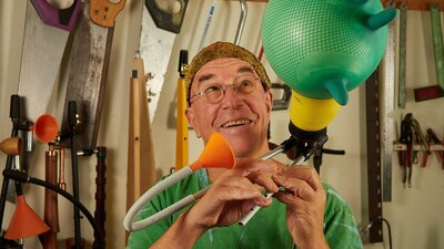 A man holds a contraption made from a funnel, a pipe, and blown up green rubber glove.