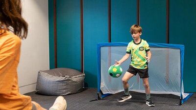 A child kicks a soccer ball towards a goal, where a child in a Matildas Jersey defends the ball.