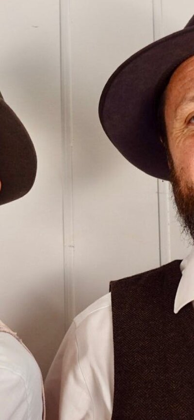 Two men in matching shirts, vests and hats smiling at camera
