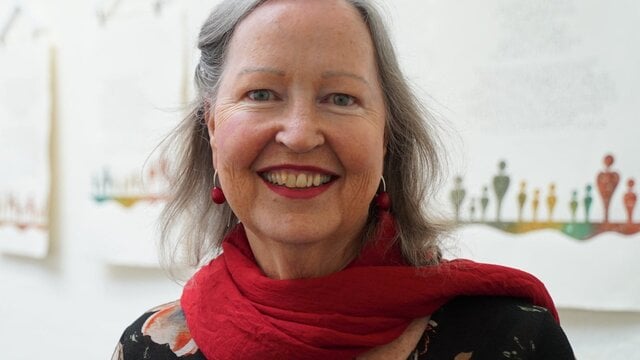 Gemma Black A smiling woman with gray hair wearing a red scarf over a dark floral top.