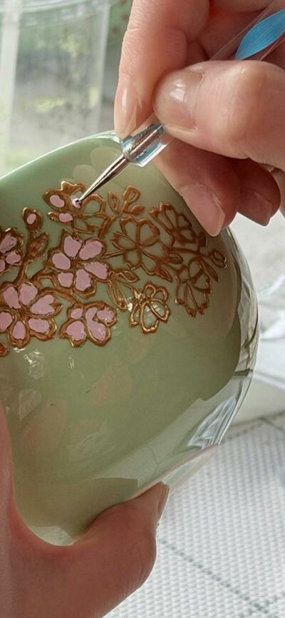 A guest paints cherry blossoms on a ceramic matcha bowl during the workshop Close-up of hands painting pink and gold flowers on a green ceramic matcha bowl