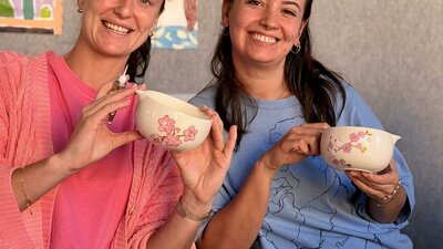 Two smiling women holding ceramic matcha bowls they painted with pink cherry blossoms