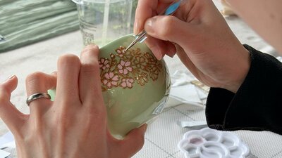 Close-up of hands painting pink and gold flowers on a green ceramic matcha bowl