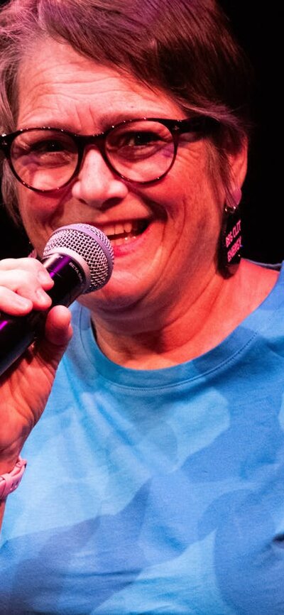 Comedian Sarah Stewart Woman wearing glasses and blue teeshirt holding mocrophone and smiling