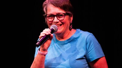 Woman wearing glasses and blue teeshirt holding mocrophone and smiling