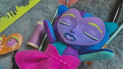 Colourful handmade plush face surrounded by thread, pens, and soft craft materials.