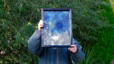 The artist holds a framed artwork of an etched mirror in front of their face, blocking it.
