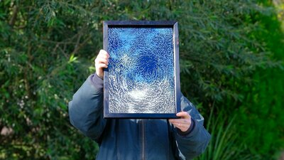 The artist holds a framed artwork of an etched mirror in front of their face, blocking it.