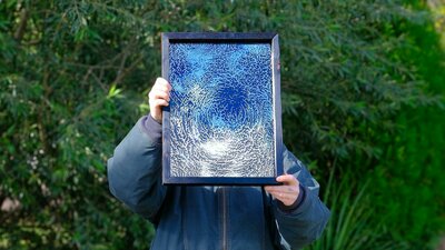April Widdup in their studio The artist holds a framed artwork of an etched mirror in front of their face, blocking it.