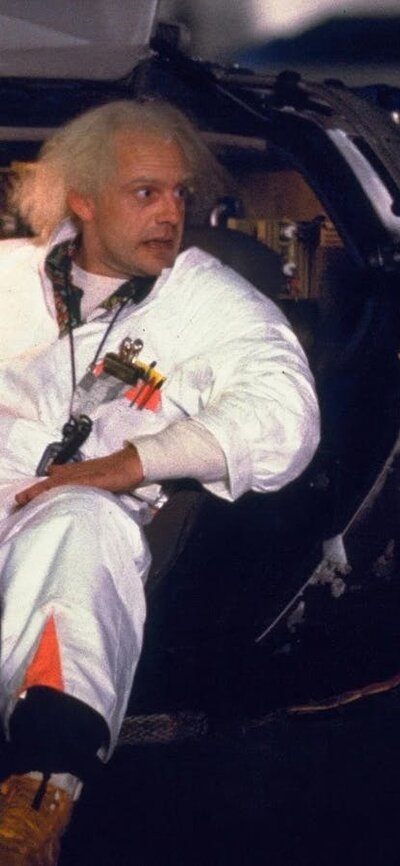 Two men facing each other, man on left sitting in car in white doctors suit