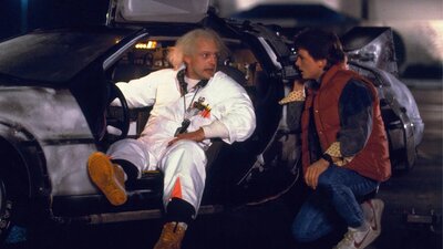 Two men facing each other, man on left sitting in car in white doctors suit