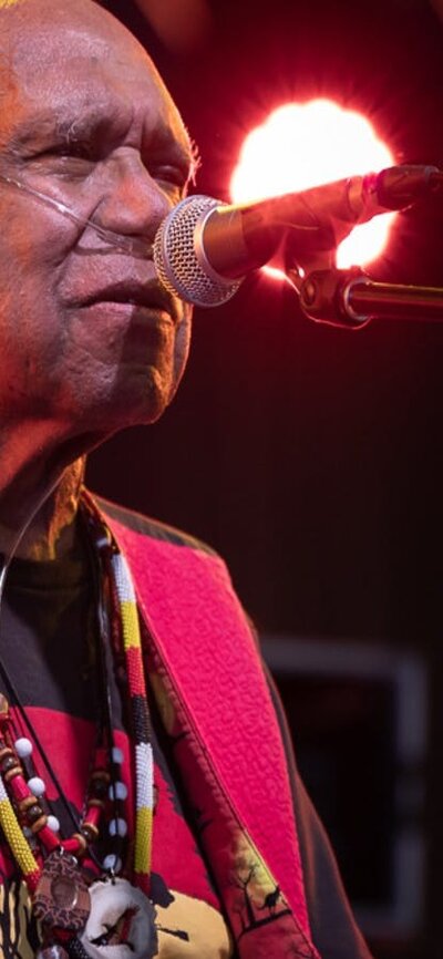 Uncle Archie roach sings on stage, wearing a black shirt and red waistcoat