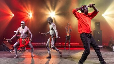 3 members of Yothu Yindi perform. There are 2 dancers in the background in traditional dress