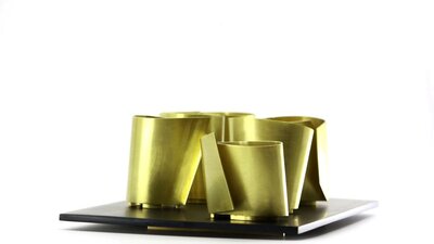 A collection of brass metal vessels with sharp angles sits on a platter against a white background