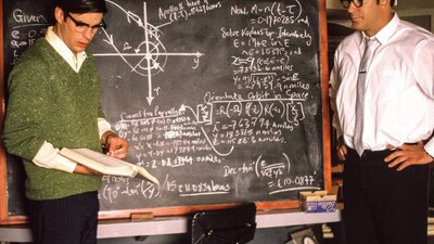 Two men stand with a blackboard with calculations on it