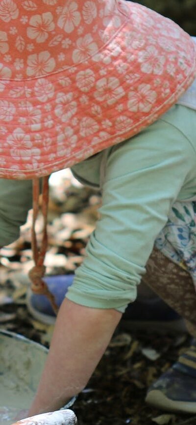 A young child wearing a sunhat, mixing dirt and water in an old frying pan